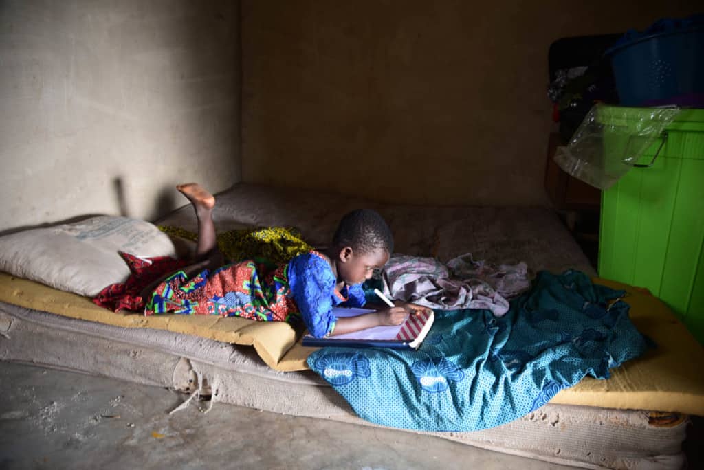 Girl asper, wearing a white shirt and gray and white striped shorts, is sitting on his bed at home and he is using a cell phone with his feet. In the foreground of the picture is a fan. Behind his is a blue and white sheet hanging up as a room separator.