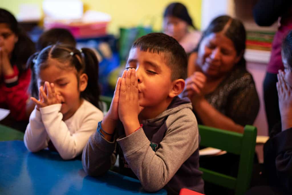 A group of women and children are sitting in a classroom praying. Their eyes are closed and their hands are together in front of their faces. The tables and chairs in the room are green and blue.