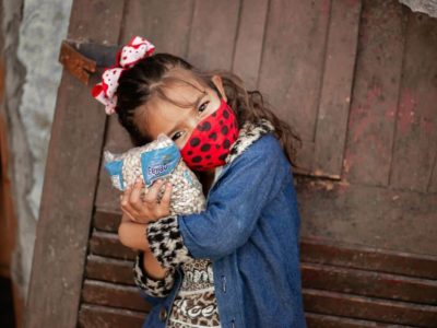 Débora is wearing a yellow shirt with a black pattern on it and a blue jacket. She is also wearing a red and black face mask. She is hugging a bag of beans outside her house.