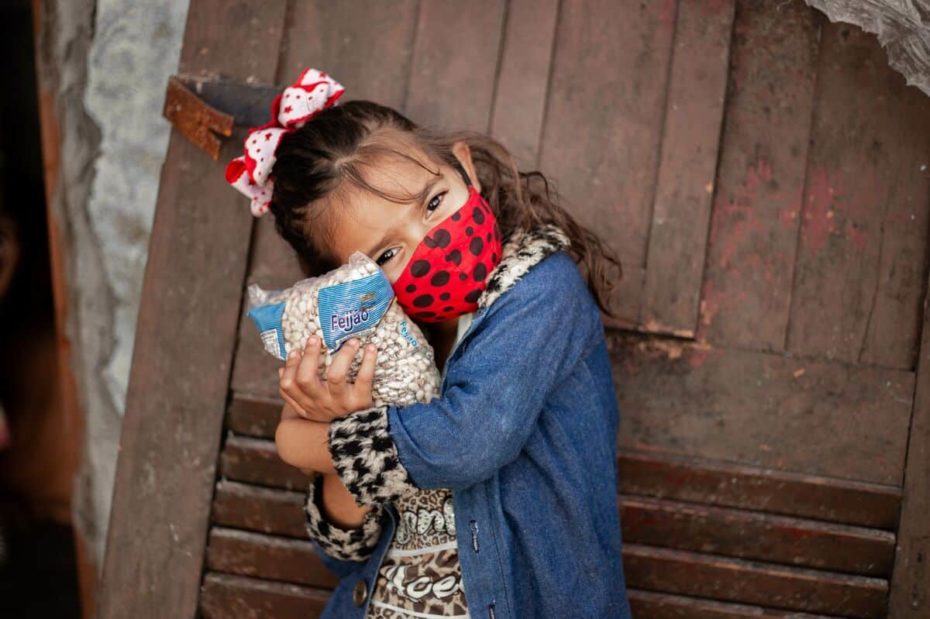 Débora is wearing a yellow shirt with a black pattern on it and a blue jacket. She is also wearing a red and black face mask. She is hugging a bag of beans outside her house.