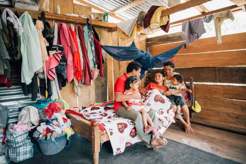 Enan with his parents and two of his siblings in his bed. The family is sitting on Enan's bed together. There are clothes hanging on the wall behind them.