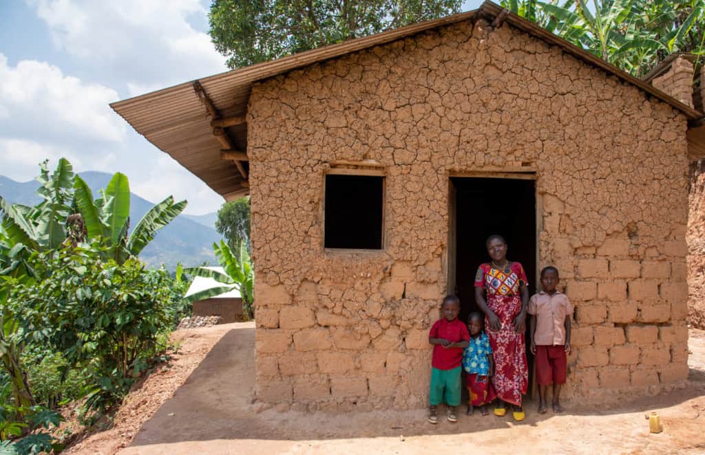 Janvier (in a red shirt and green shorts) is standing at the entrance to his mud home with with his mother and his siblings.