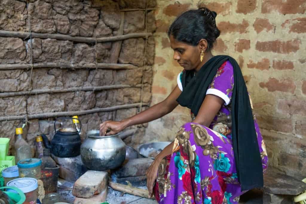 Fausiya is wearing a purple dress with a floral print and a black scarf. She is kneeling down and is cooking in her kitchen.