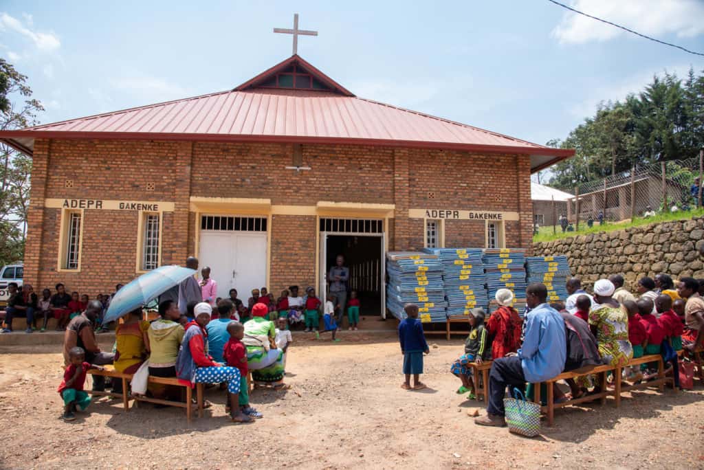 Caregivers and children are sitting on benches outside the brick church waiting to receive their new mattresses and uniforms.