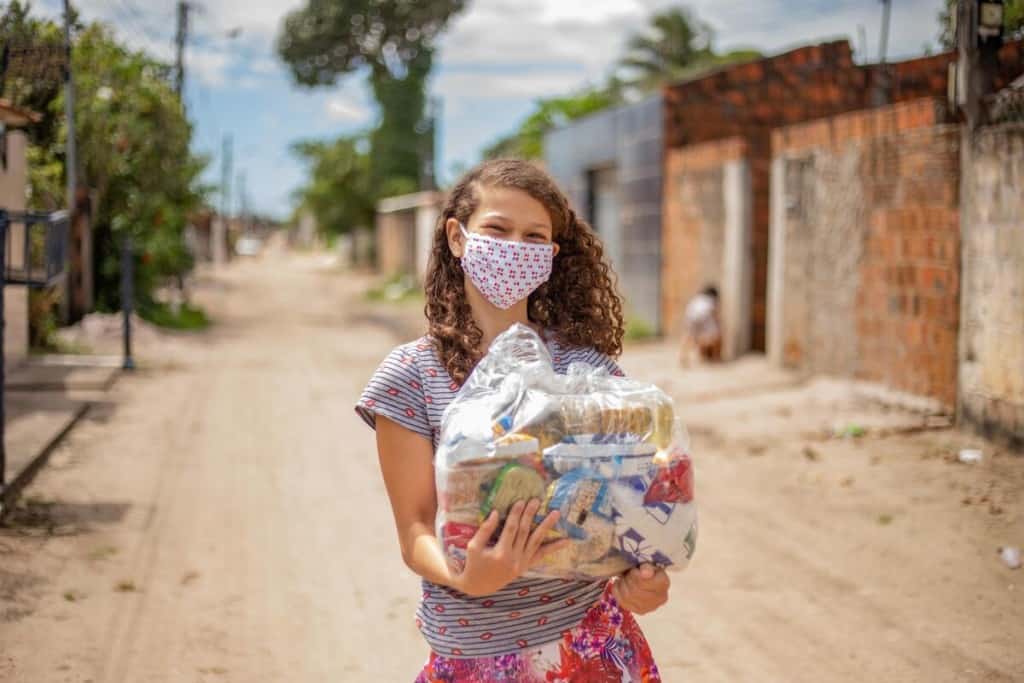 Germina is wearing a gray and black striped shirt with a red floral print skirt and a face mask. She is holding a basket of food that was given to her family by the Compassion center. She is standing on a dirt road in her neighborhood.