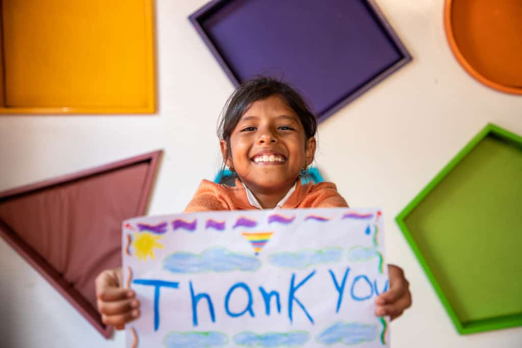 Girl wearing an orange shirt. She is standing in front of a wall with colorful shapes all over it and is holding up a piece of paper that says Thank You.