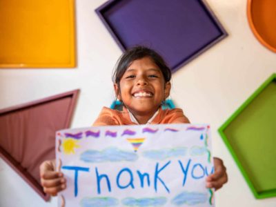 Girl wearing an orange shirt. She is standing in front of a wall with colorful shapes all over it and is holding up a piece of paper that says Thank You.