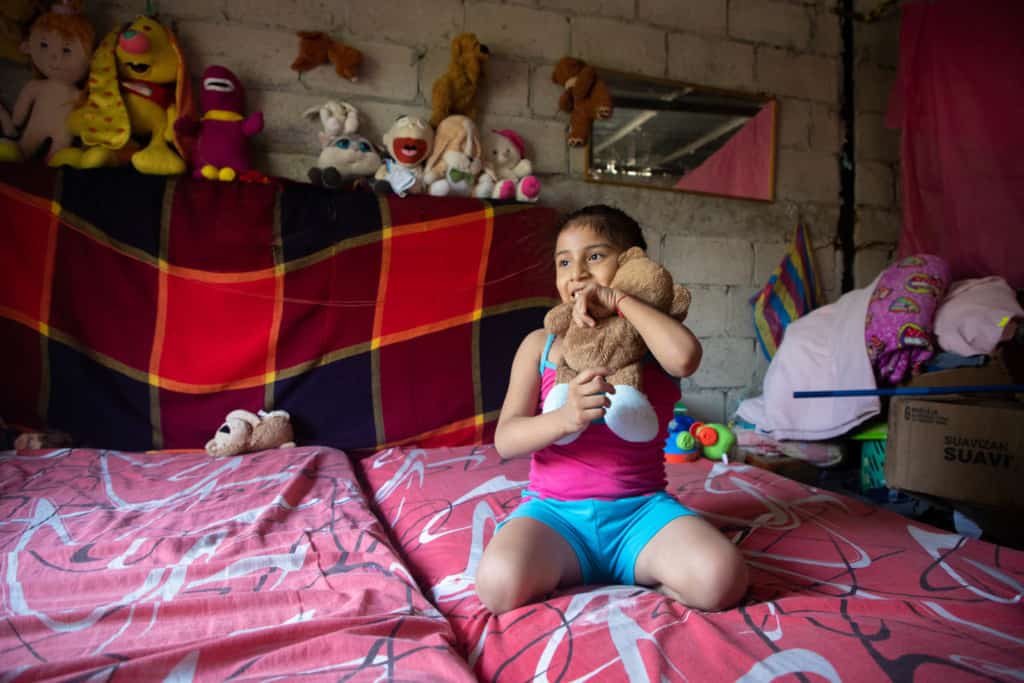 Girl wearing a pink shirt and blue shorts. She is sitting on her bed hugging her teddy bear. Behind her are her toys.