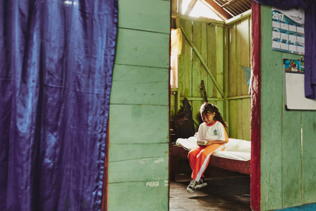 A girl in white and orange sits on a bed holding and looking at a book through a doorway with green wood walls, red trim and purple curtain.