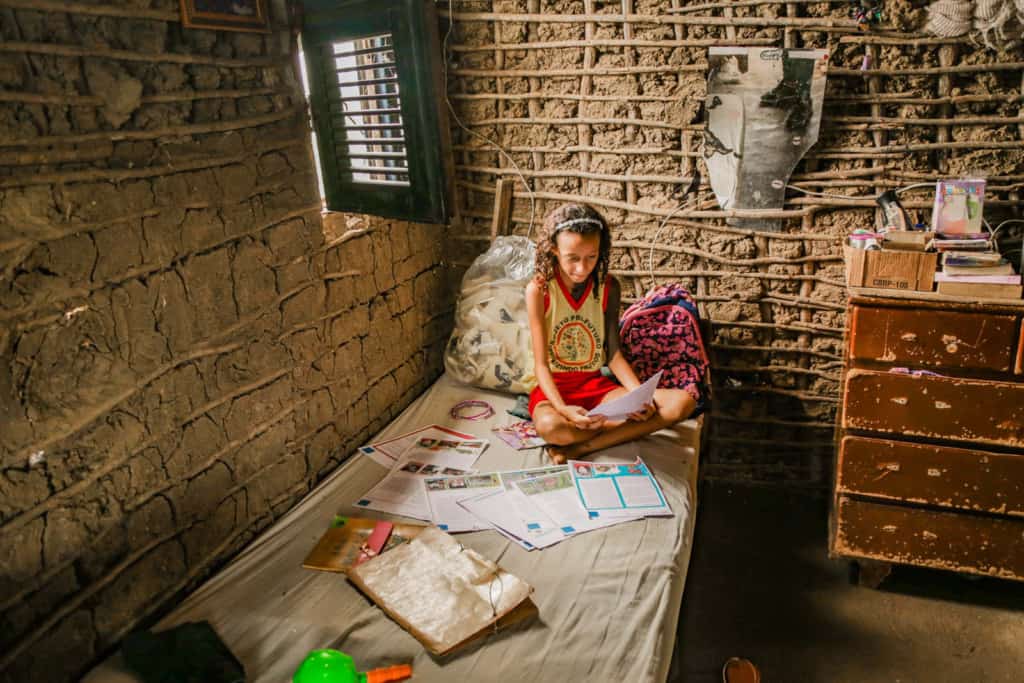 Girl reads one of her sponsor’s letters on the bed she shares with her older sister every night. Kaylane is wearing a yellow shirt and red skirt.