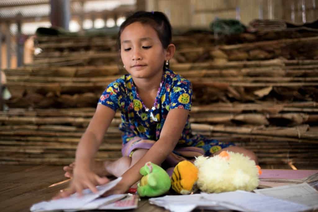 Girl wearing a floral patterned shirt. She is sitting on the floor and is looking at books. There are stuffed animals on the floor.