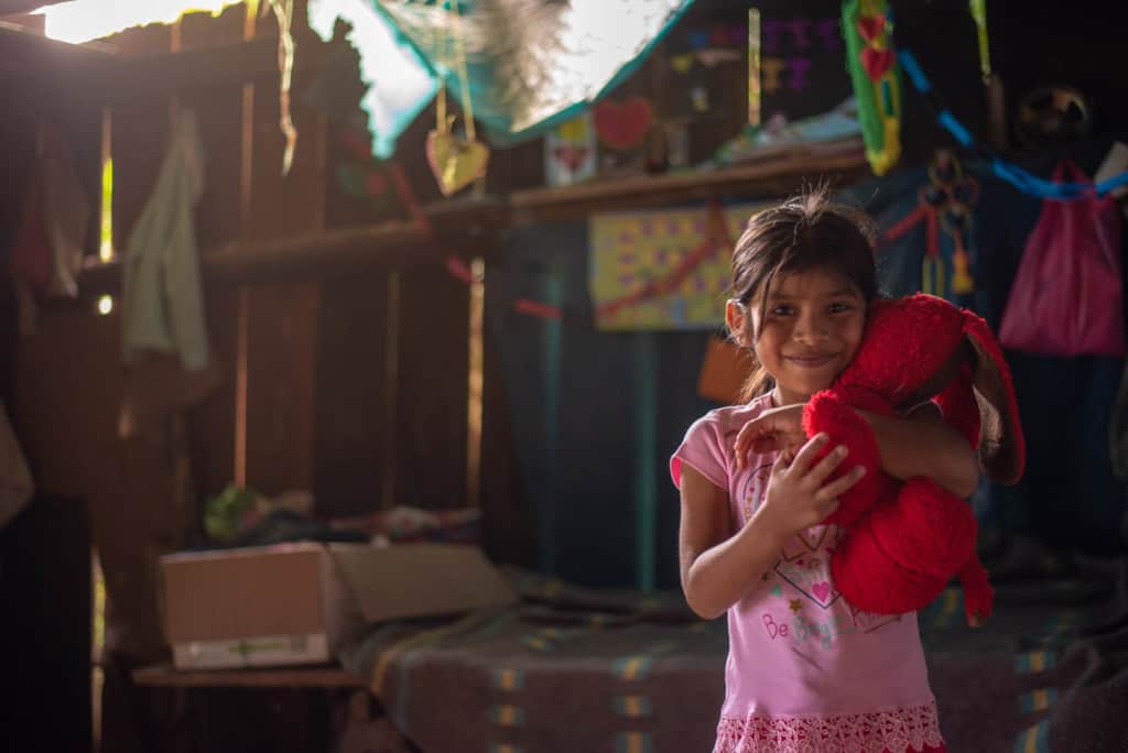 Girl holding her teddy bear in her room. She is wearing a pink tee shirt and is smiling at the camera. There is a wooden fence behind her.