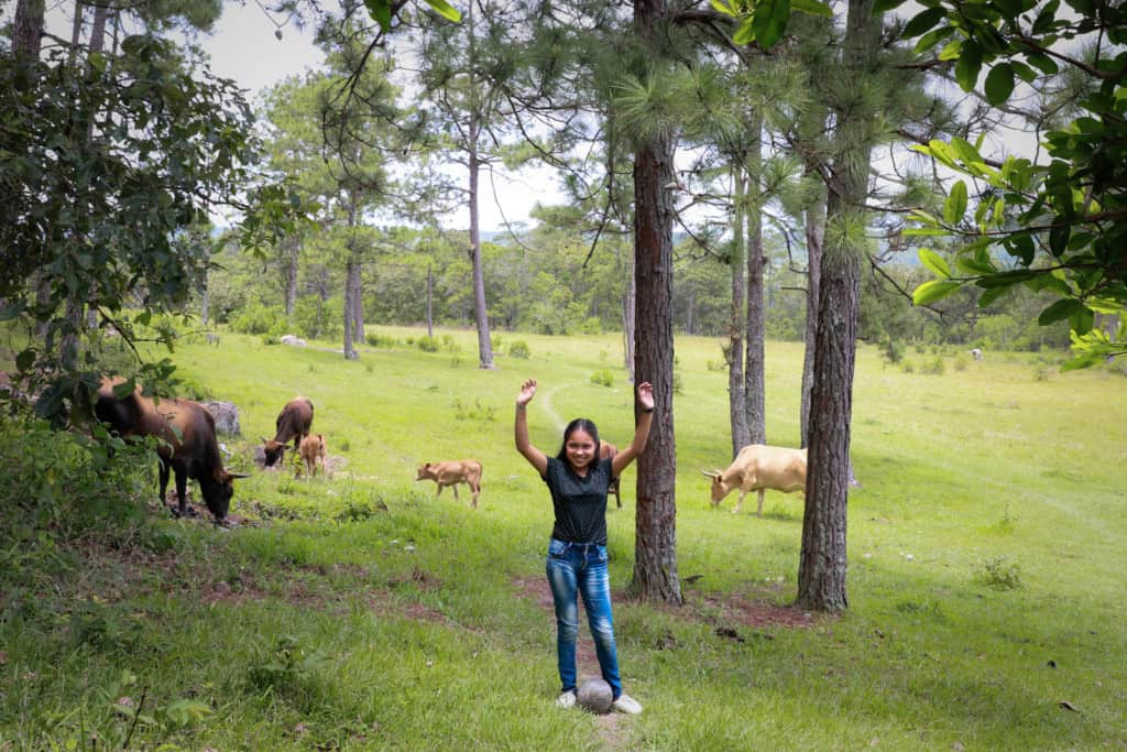Girl wearing a black blouse with little blue flowers on it. She’s also wearing jeans and white tennis shoes. She is standing in a field with cows and she is holding her hands in the air.