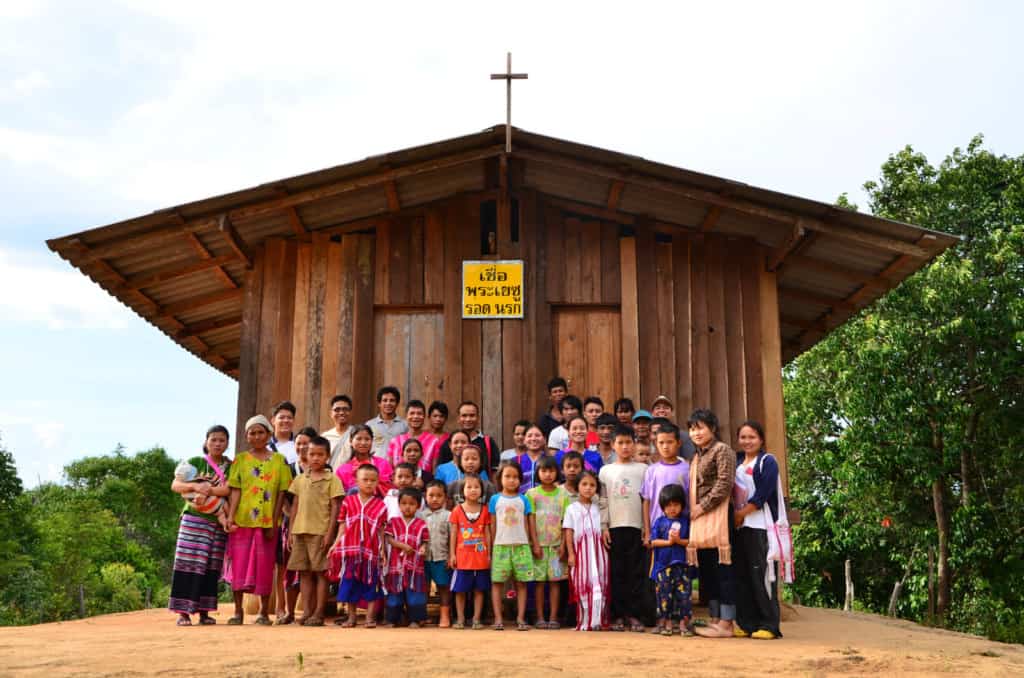 New students and parents pose outside of a wooden church building with a cross. They are standing in the dirt and there are trees in the background.