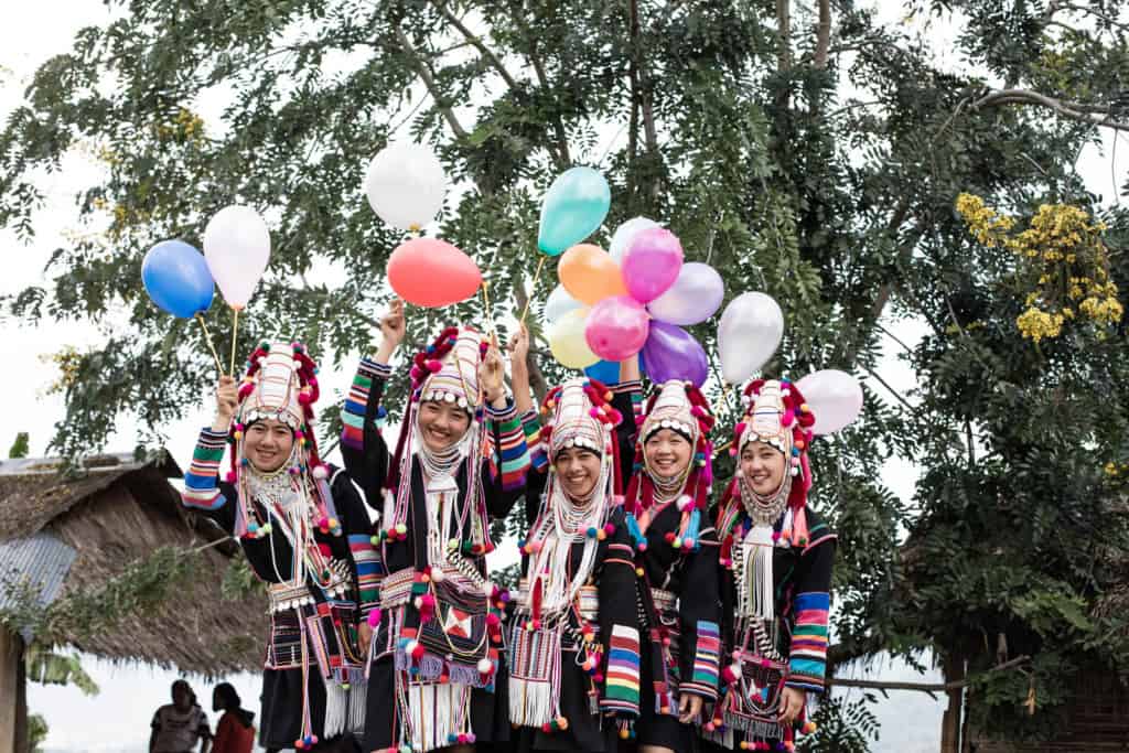 A group of women standing on the back of a truck, wearing their Akha traditional custom dress and hats, holding colorful balloons and smiling.