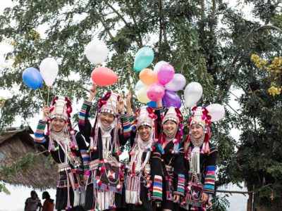 A group of women standing on the back of a truck, wearing their Akha traditional custom dress and hats, holding colorful balloons and smiling.