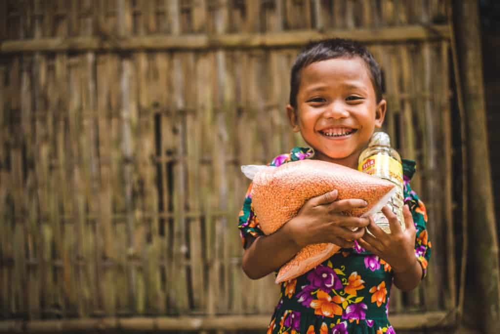 Girl wearing a green dress with a floral print. She is standing in front of a bamboo wall and is holding food she received from the Compassion center.