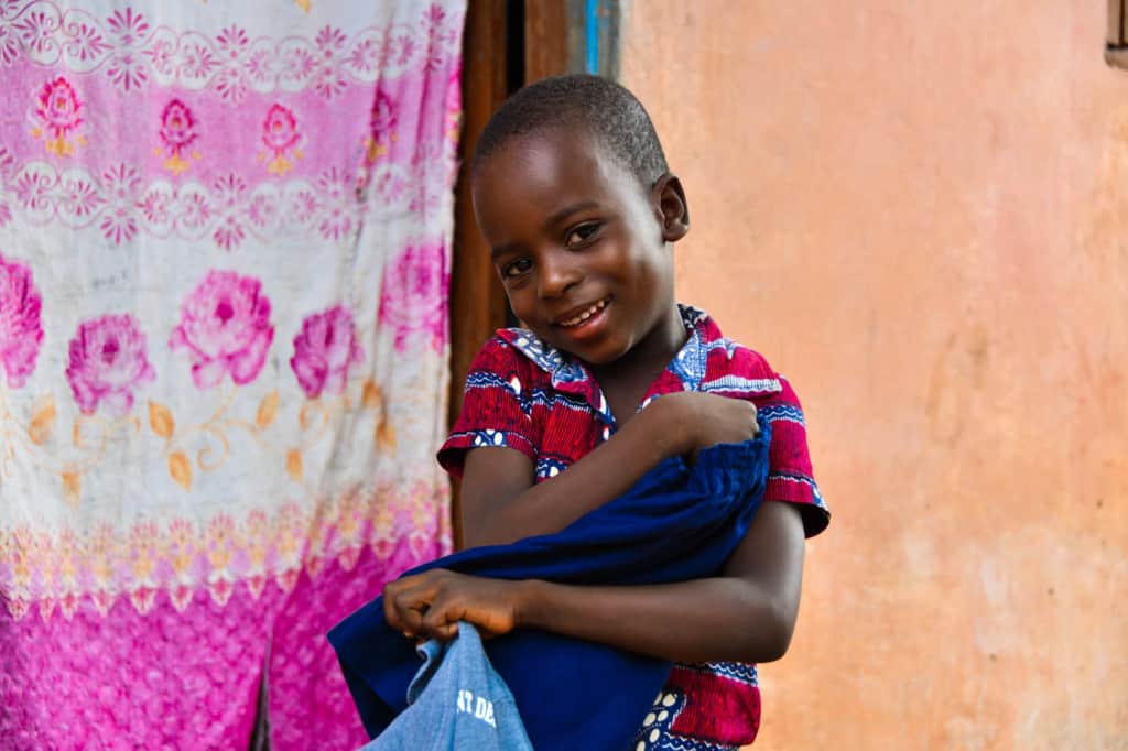 Herve is wearing a red shirt with white and blue patterns on it. He is standing in front of his hom, which is tan with a pink floral print sheet hanging in the doorway. Herve is holding clothes he received as a gift last Christmas.