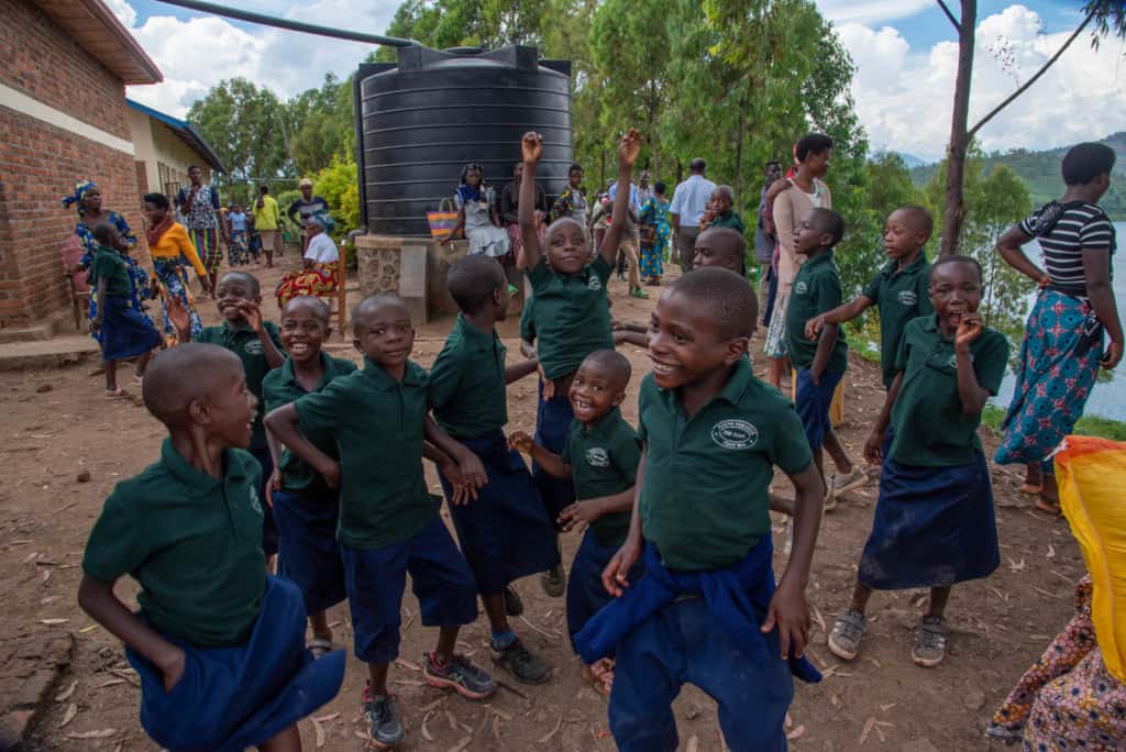 A group of children in green and blue uniforms are outside the project playing. There are adults standing around in the background.