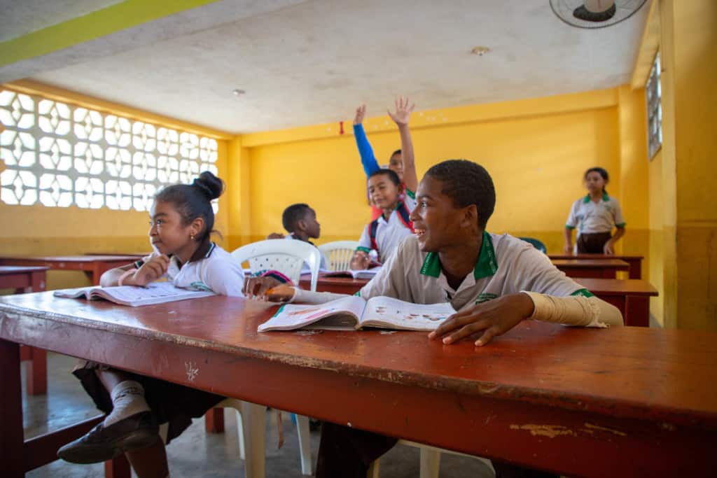 Steven (front right) and other children wearing white, green and black uniforms are sitting at long red desks, tables in a classroom with yellow walls. There are books on the tables in front of them. Steven is smiling and looking to the side. Two children in the background are raising their hands.