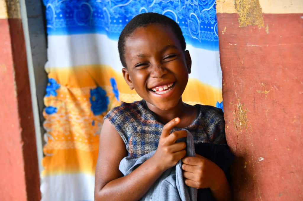 Esther is standing in the doorway of her home and is holding a dress she received as a gift last Christmas. There is a blue, white, and orange curtain in the doorway.