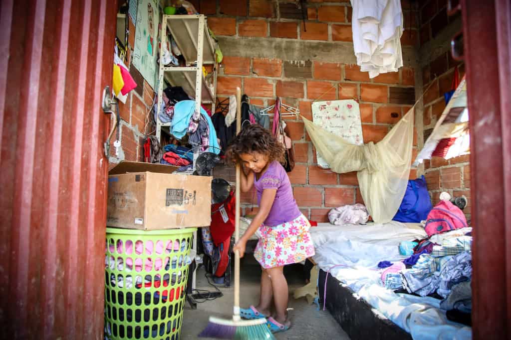 A girl in a purple shirt is standing inside her home holding a broom and sweeping. The walls are brick and she is next to two beds with clothes on them.