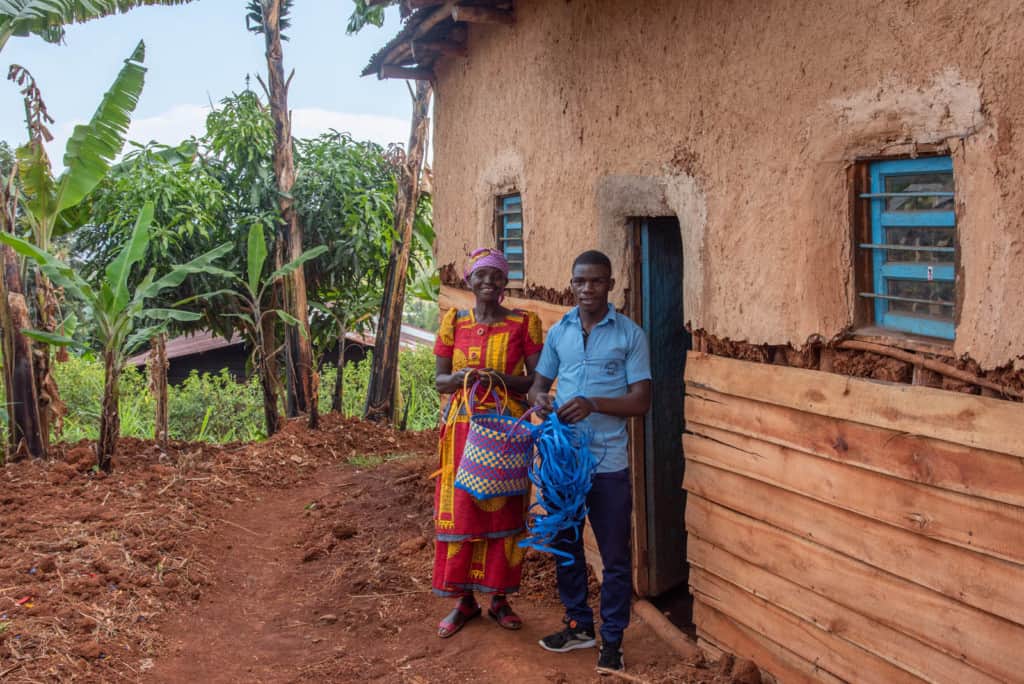 Sebastian is wearing dark blue pants and a blue shirt. He is standing outside his home with his mother, Belancille, wearing a red and yellow dress with a maroon head covering. They are showing the baskets they have weaved. Their home is wooden on the bottom and cement on the top half. There are trees in the background.