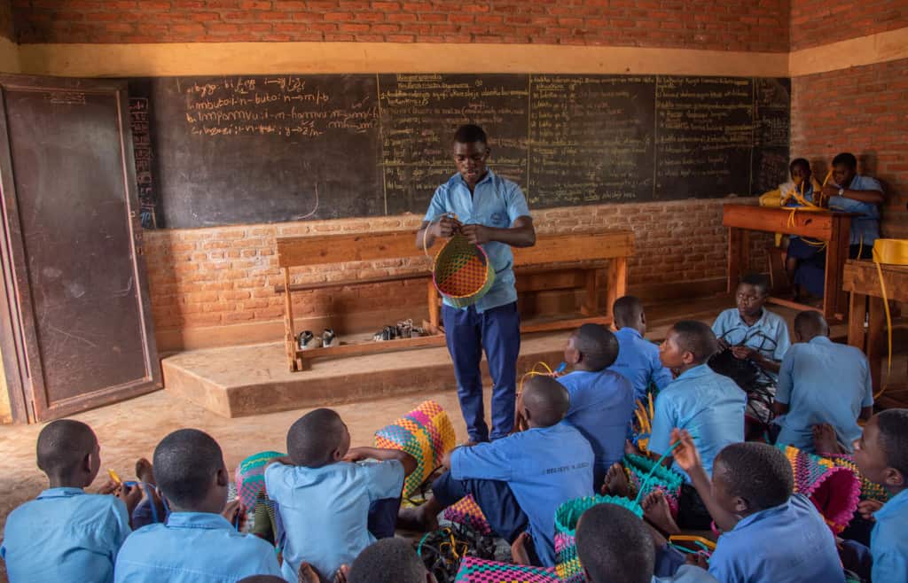 Sebastian is wearing dark blue pants and a blue shirt. He is teaching youth at the Compassion center how to weave bags in one of the classrooms. Behind him is a brick wall with a large black board on it.