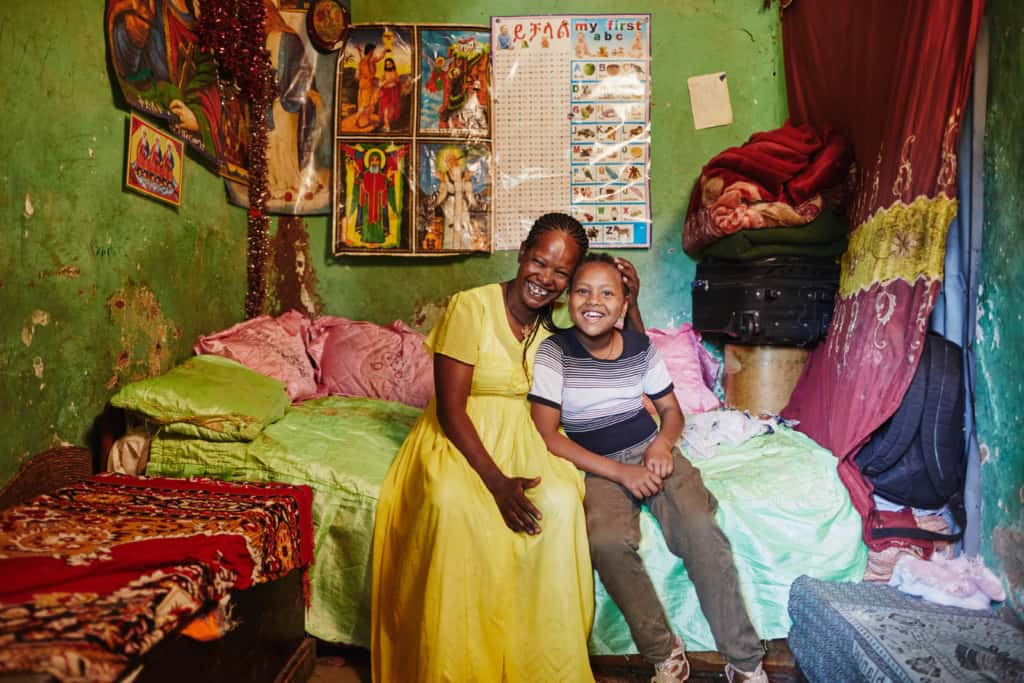 A mother, in a yellow dress, is sitting on a bed with her daughter with their heads pressed together. The walls are green and there is a red curtain, blanket and table cloth.