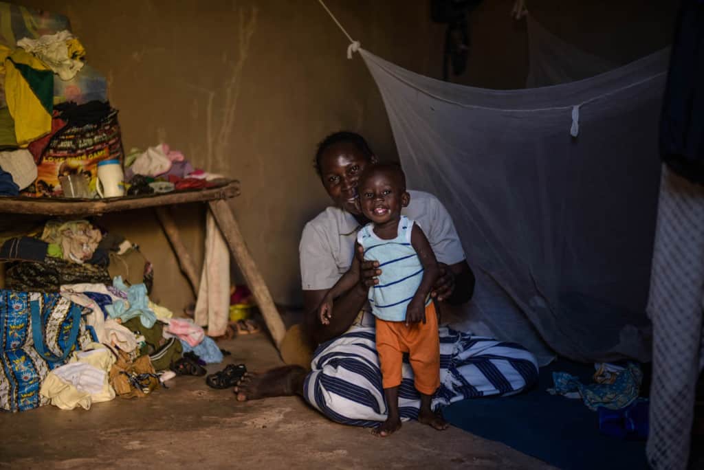 A mother and her toddler are siiting inside their home. On the left is a wooden table with clothes and other household belongings. Behind them is a mosquito net hanging from the wall.