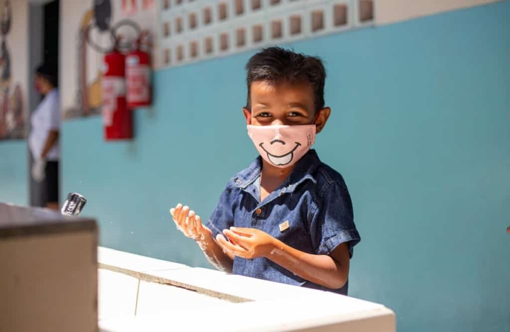 Pedro is wearing a denim shirt with a pink face mask. He is washing his hands in the sink. The wall behind him is blue.