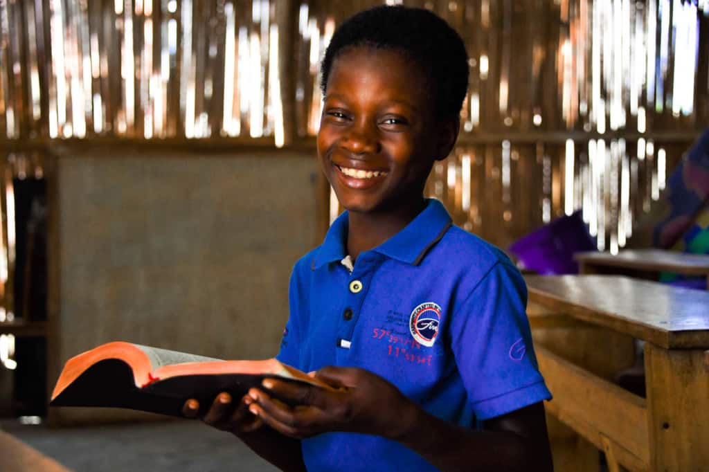 Deborah is wearing a blue shirt. She is sitting inside the Compassion center and is holding a Bible. She is sitting at a wooden table.