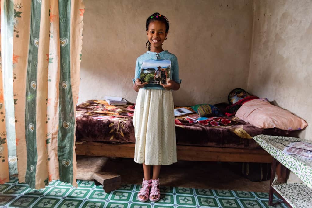 Girl wearing a blue shirt and white skirt. She is standing in her home in front of her bed, holding pictures of her sponsors.