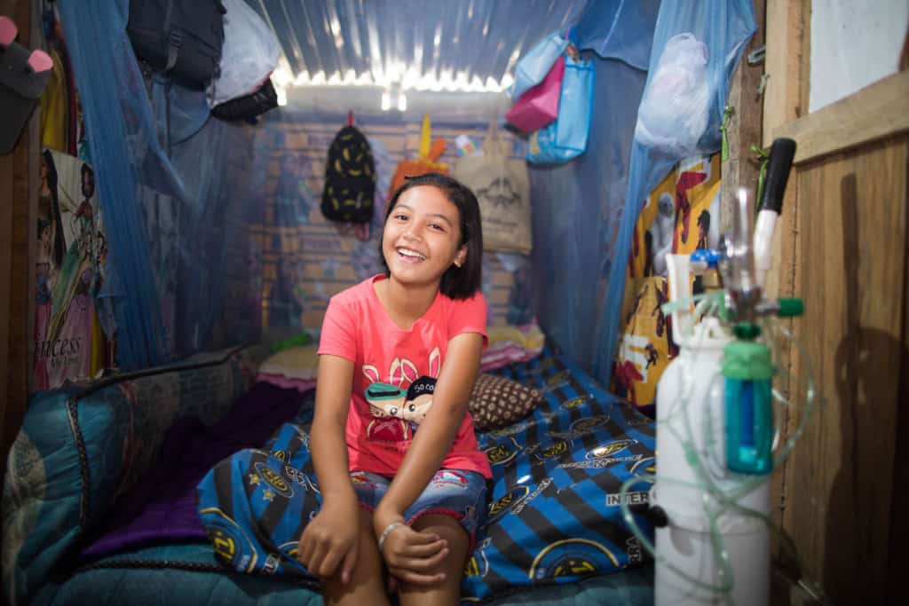 Girl wearing shorts and a red shirt. She is sitting on her bed. There is an oxygen tank beside her bed.