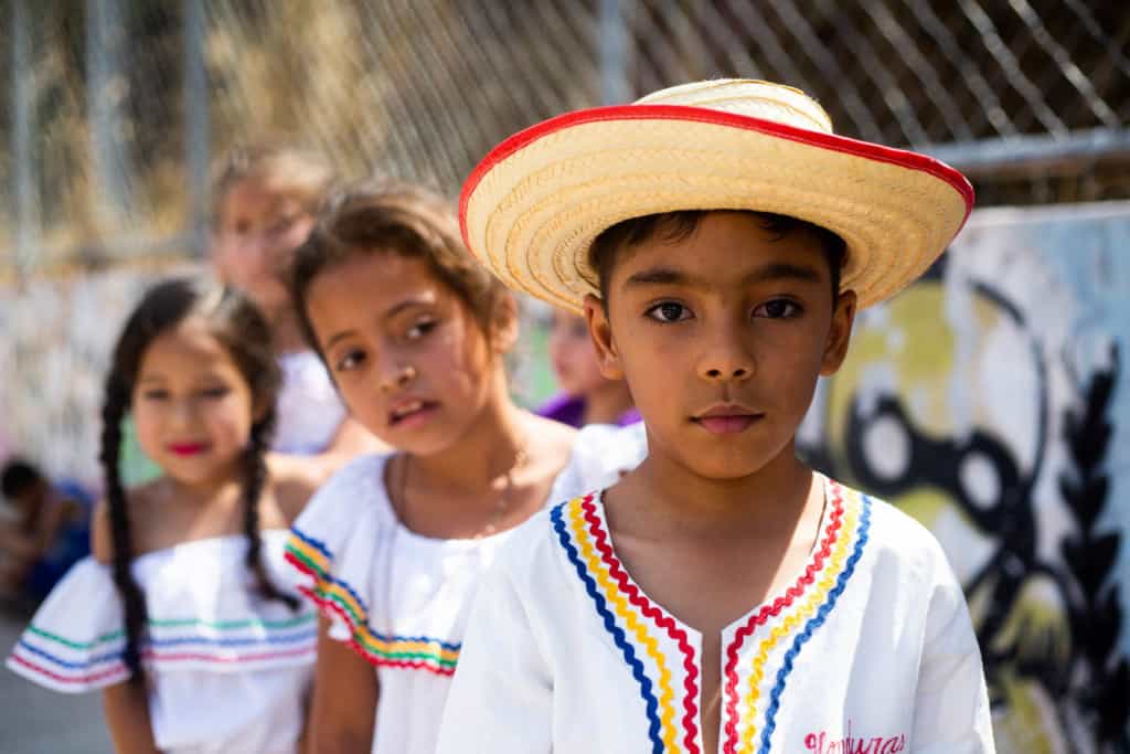 A boy in a white shirt and a straw hat with red trim is standing in front of two girls next to a chain link fence.