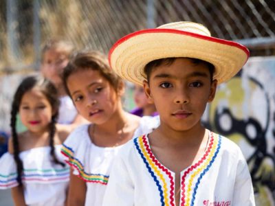 A boy in a white shirt and a straw hat with red trim is standing in front of two girls next to a chain link fence.