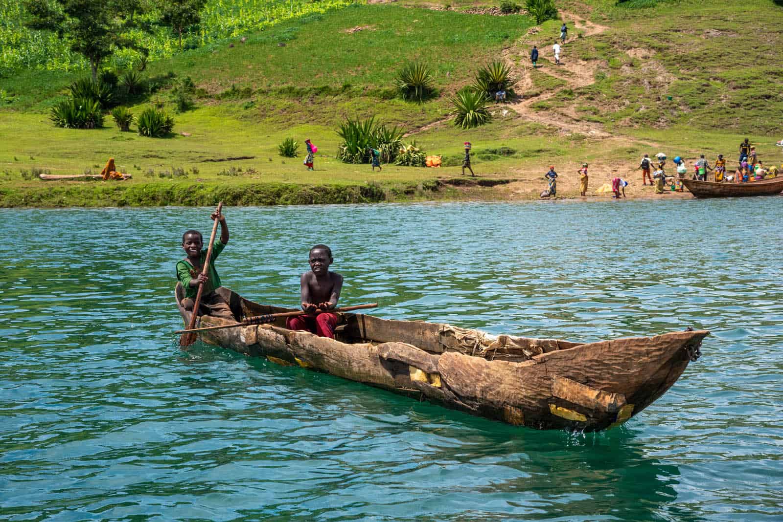 Two children are in a wood canoe on a lake. There are more people unloading items from another boat in the background.