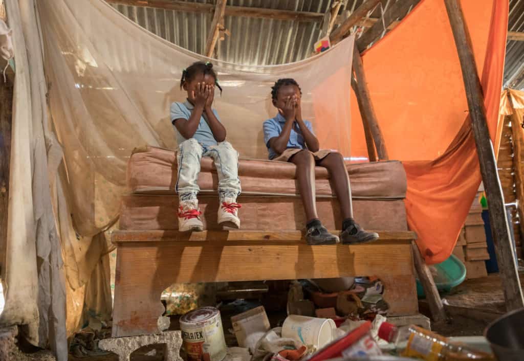 Two sisters sitting on their bed that has a mosquito net on it. There is an orange curtain to divide their room from the dining room. The bed is on top of a concrete block to give it height and protect the girls from water when the river floods.