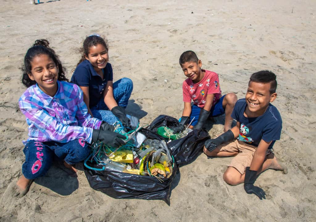 Several children are sitting on the beach picking up trash. The children are all wearing black gloves.