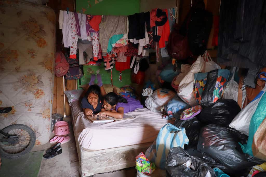 Sisters laying down on the bed they share with their grandmother. One is wearing a gray dress with pink hearts on it and purple socks. The other is wearing a purple dress.