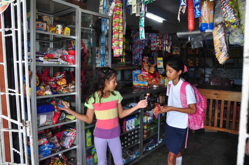 A 9 year old girl wearing a colorful striped shirt, stands beside a counter market store shelf shopping for candy as she shows her friend standing beside her who is wearing a white shirt and pink backpack a lollipop in her hand. Candy and other food supplies on shelves and hang around the store all around them.