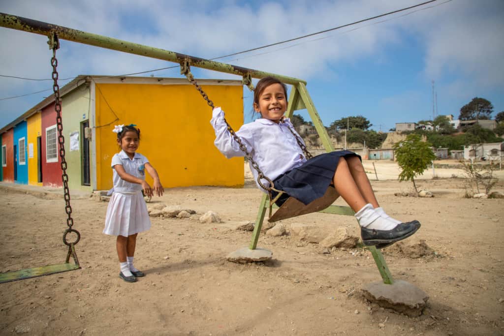 Girl wearing a white shirt and black skirt. She is sitting on a swing while her friend pushes her. The school building is behind them.