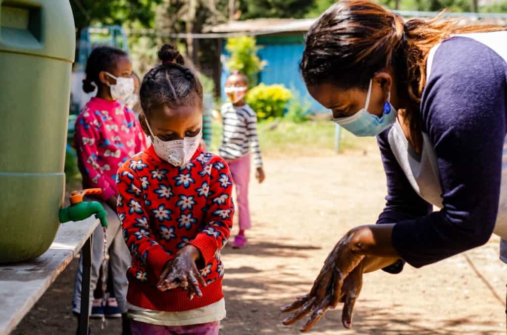 A group of children is standing in line waiting to wash their hands. A girl wearing a red sweater is washing her hands as a staff member at Compassion's partner church instructs her. All the children are wearing face masks and are maintaining proper distance from each other.