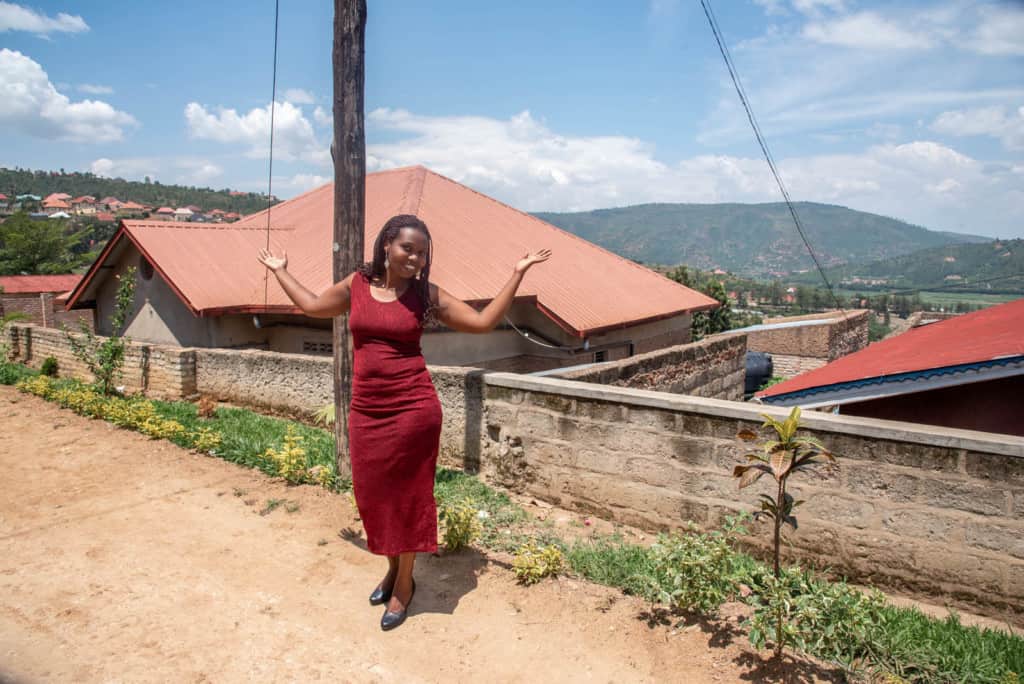 Jeannette is wearing a burgundy dress and is standing on a dirt road with houses in the background. She is posing for the camera.