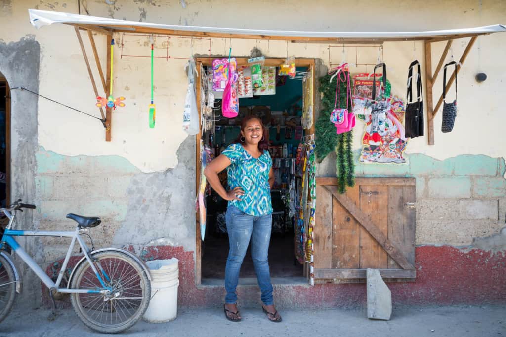 Woman wearing a blue shirt and jeans stands smiling looking out from the back of her market shop selling goods merchandise food snacks in a small building.
