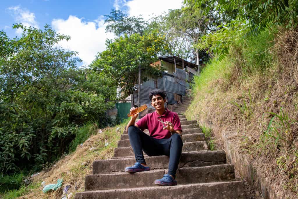 Miguel, in a red shirt and black pants, is outside sitting on a steep set of stairs. He is holding a pair of scissors and a comb.