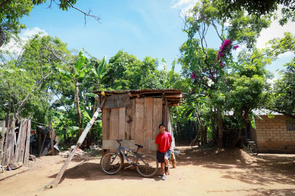 Boy wearing a red shirt and shorts. He is leaning against a wooden barn. His bicycle is next to him.