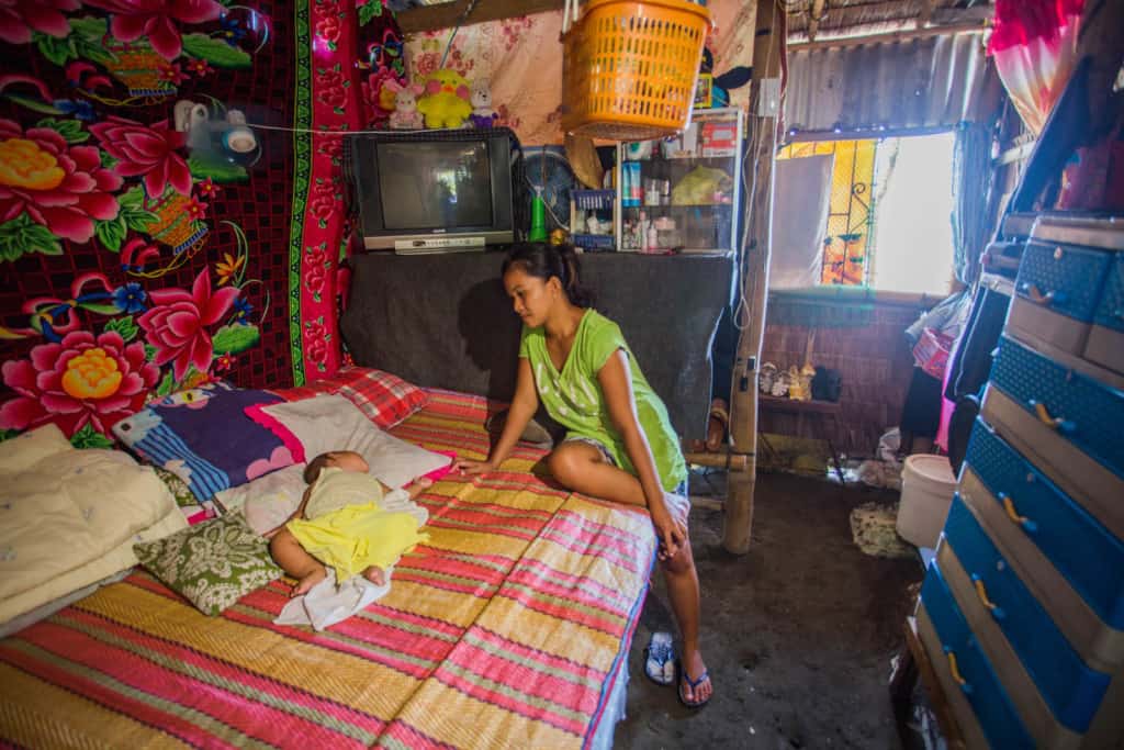 A mother wearing a green shirt, is sitting on a bed in her home looking at her baby who is sleeping. The house is very crowded.