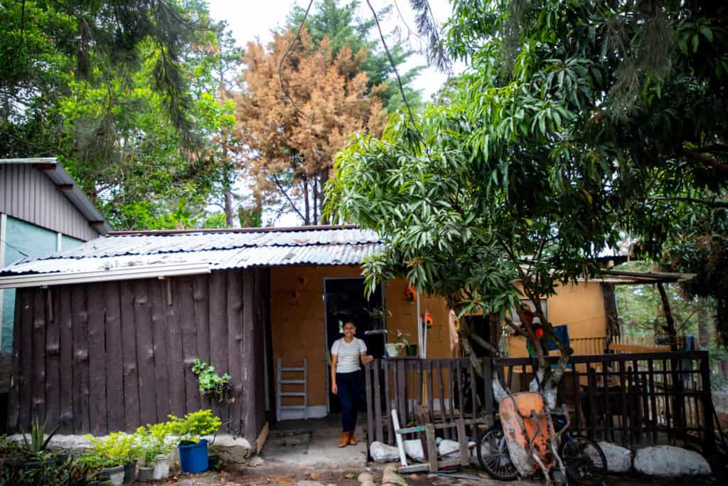 Young woman wearing a black and white striped shirt and jeans. She is standing in front of her home, which is made of wood with a corrugated metal roof. There are trees behind the house.