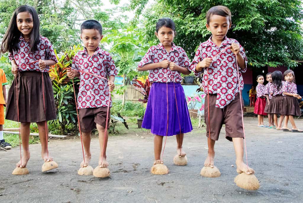 Four children in matching button-up shirts with brightly colored geoemetric patterns are playing a game where they walk on halved coconut shells, which are attached to ropes that they are holding with their hands.